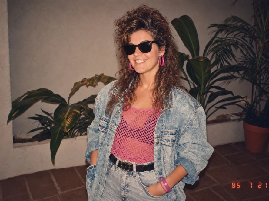 A smiling woman in black sunglasses, bright pink hoop earrings, a pink mesh top, and an oversized denim jacket poses in front of indoor plants. A red-orange date stamp reads “85 7 21” in the lower-right corner.
