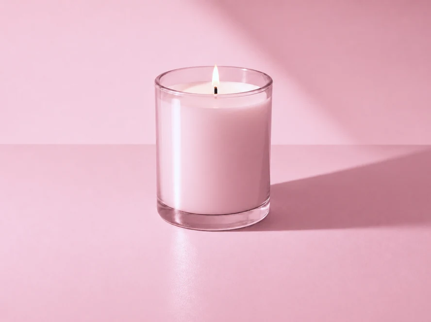 Lit candle in a pink glass jar on a pink surface, casting a long shadow.